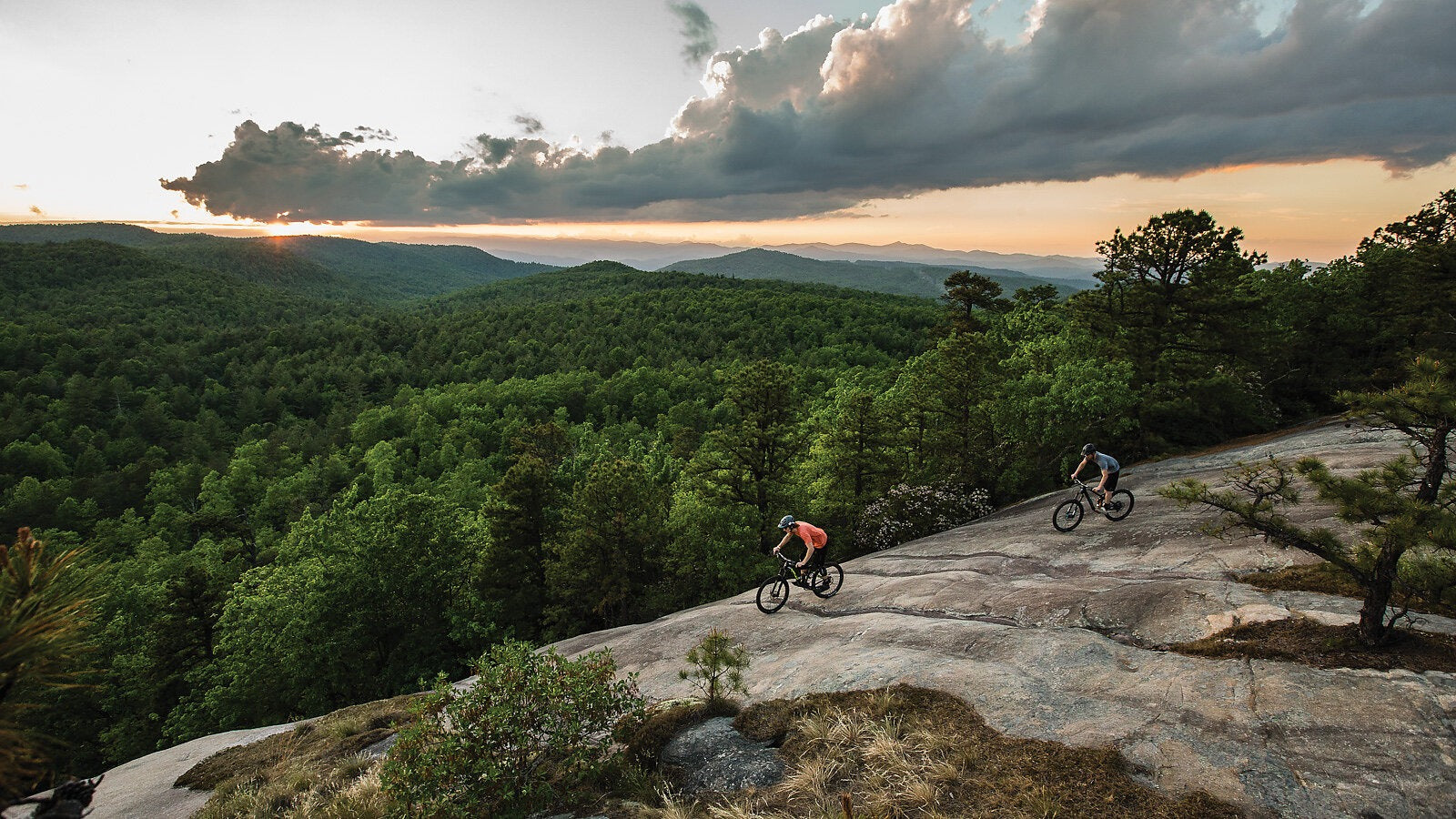 Two mountain bikers on a rocky trail with a scenic forest and sunset in the background