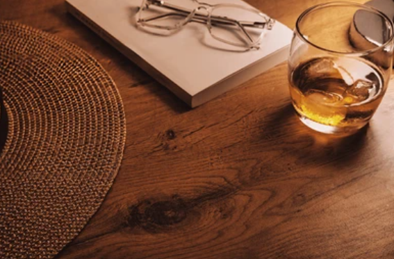 Glass of amber liquid with ice cubes on a wooden table, next to a book and glasses.